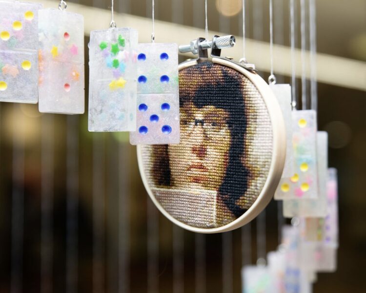 A detail shot of an art installation mobile featuring strings of dominoes with rainbow dots, and a small circular cross-stitch portrait framed in an embroidery hoop hung in between. The image is a photorealistic portrait of a young person with shoulder-length dark brown hair and glasses, wearing a somber expression. 
