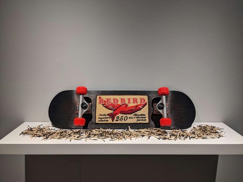 Image of a skateboard mounted horizontally on a white shelf against a gray wall. The skateboard has red wheels and a central beaded graphic resembling a vintage matchbox label that reads "Redbird" with an image of a red bird in flight. The shelf is scattered with numerous used matchsticks.